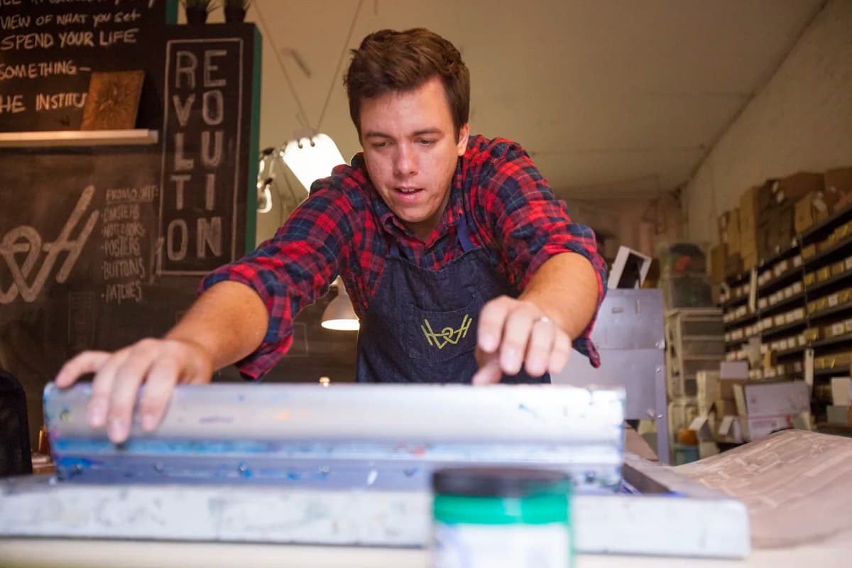 Tj pulling a screen print at the studio bench in front of a Revolution chalkboard.