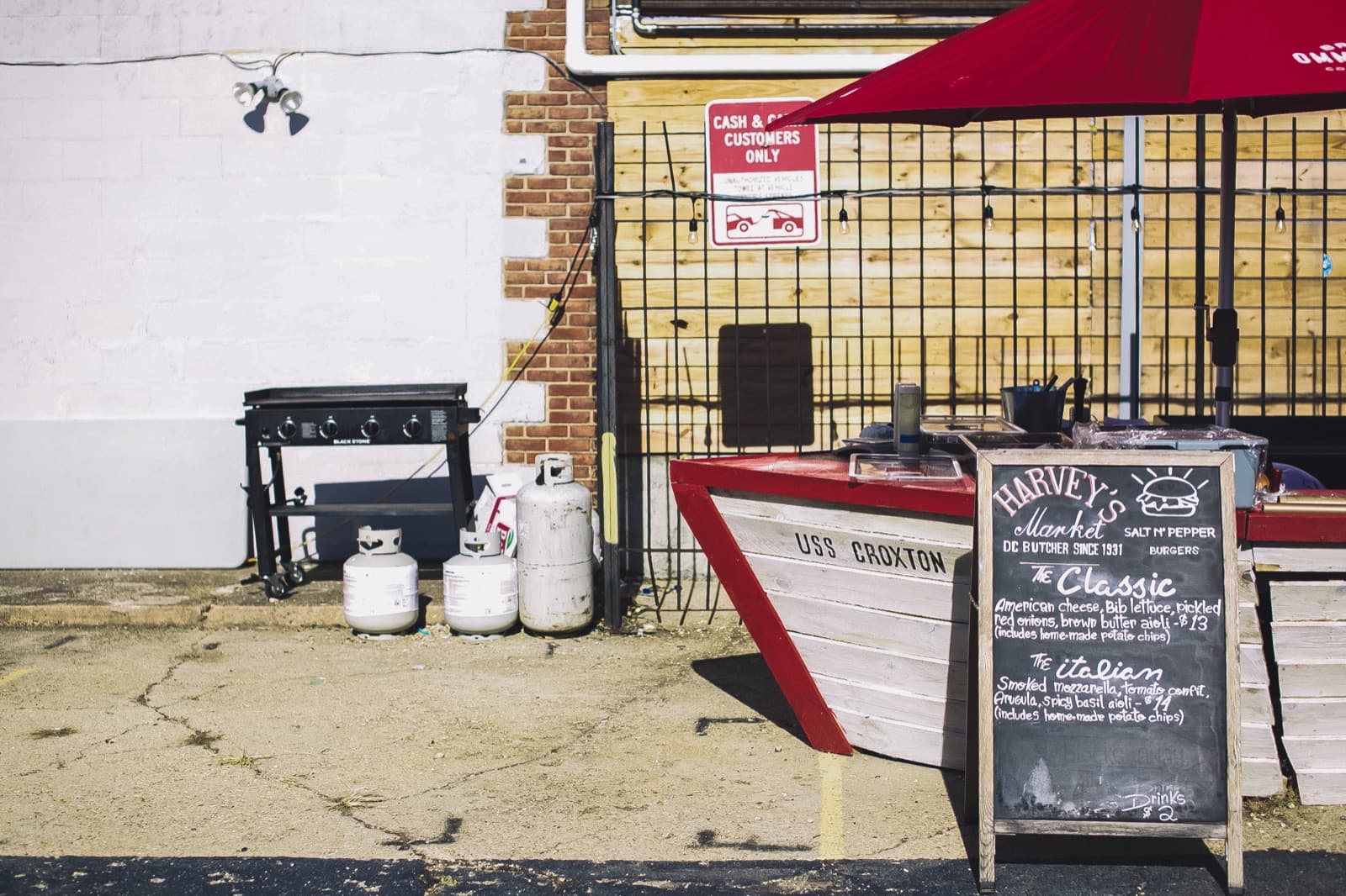 A wooden boat-shaped food stand at a DC Design Week closing party with a chalkboard menu reading Harvey’s Market and USS Croxton.