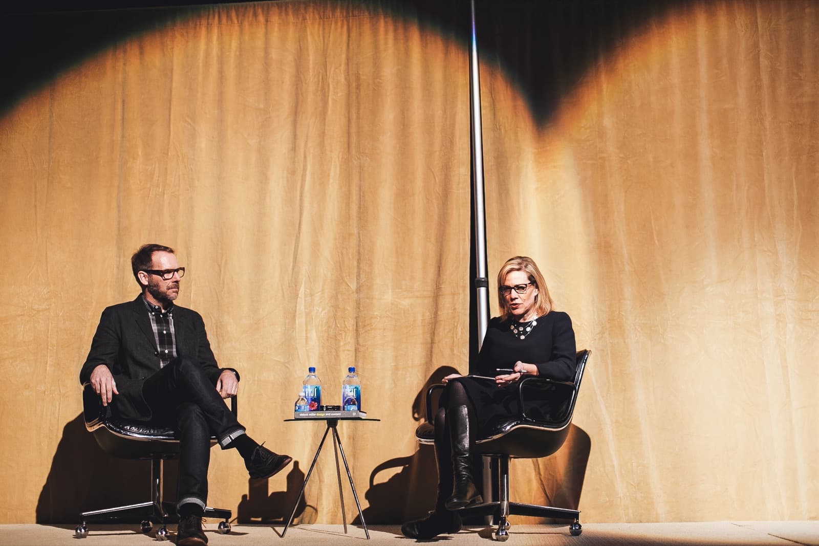 Two panelists seated against a giant amber spotlit drape, design conference stage.