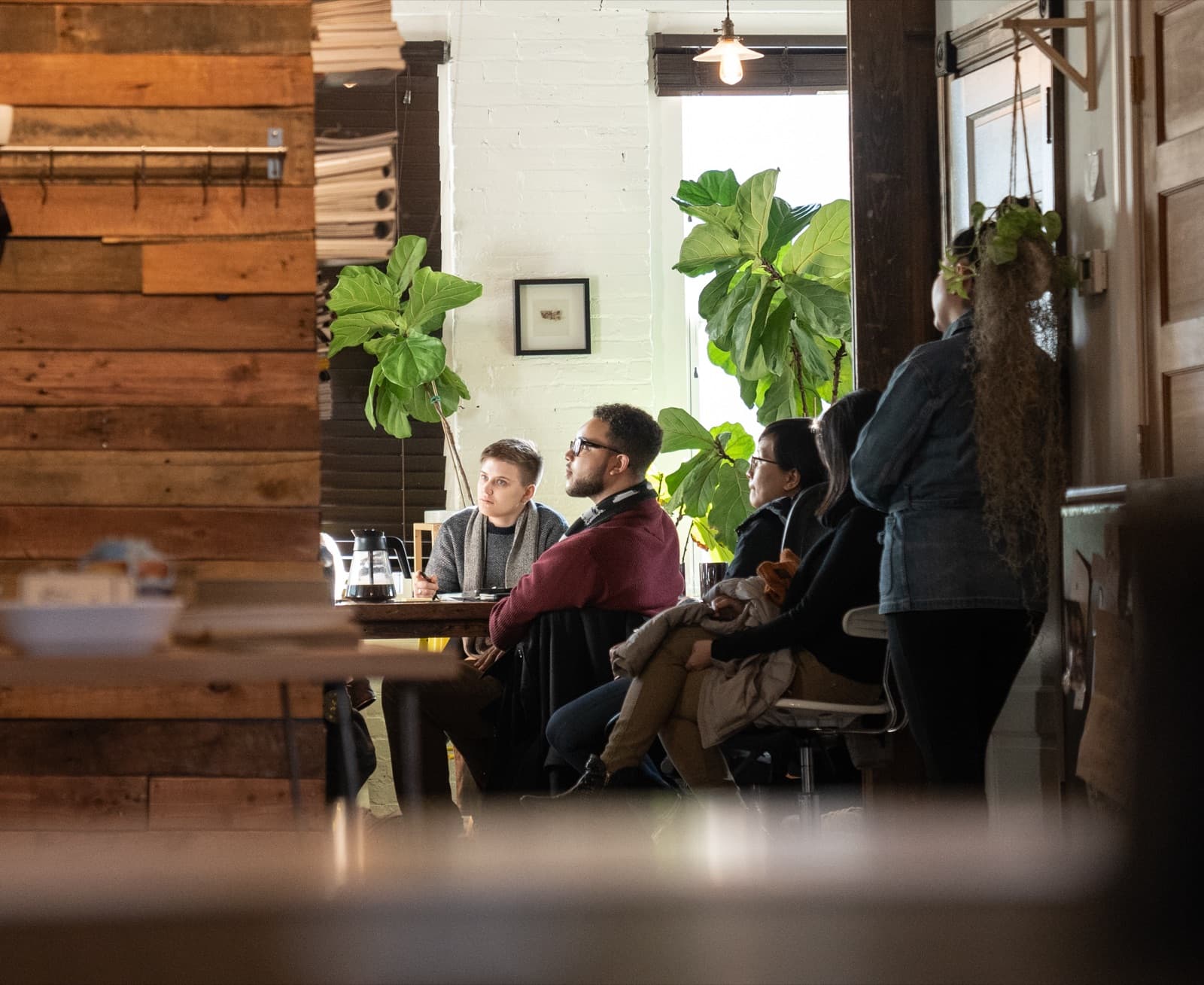 View through a reclaimed-wood doorway into the talk room, audience listening, fiddle leaf fig and hanging plants framing.