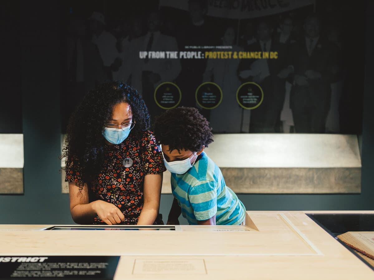 MLK Memorial Library exhibition, visitors at a maple reading table beneath the Up From the People masthead.