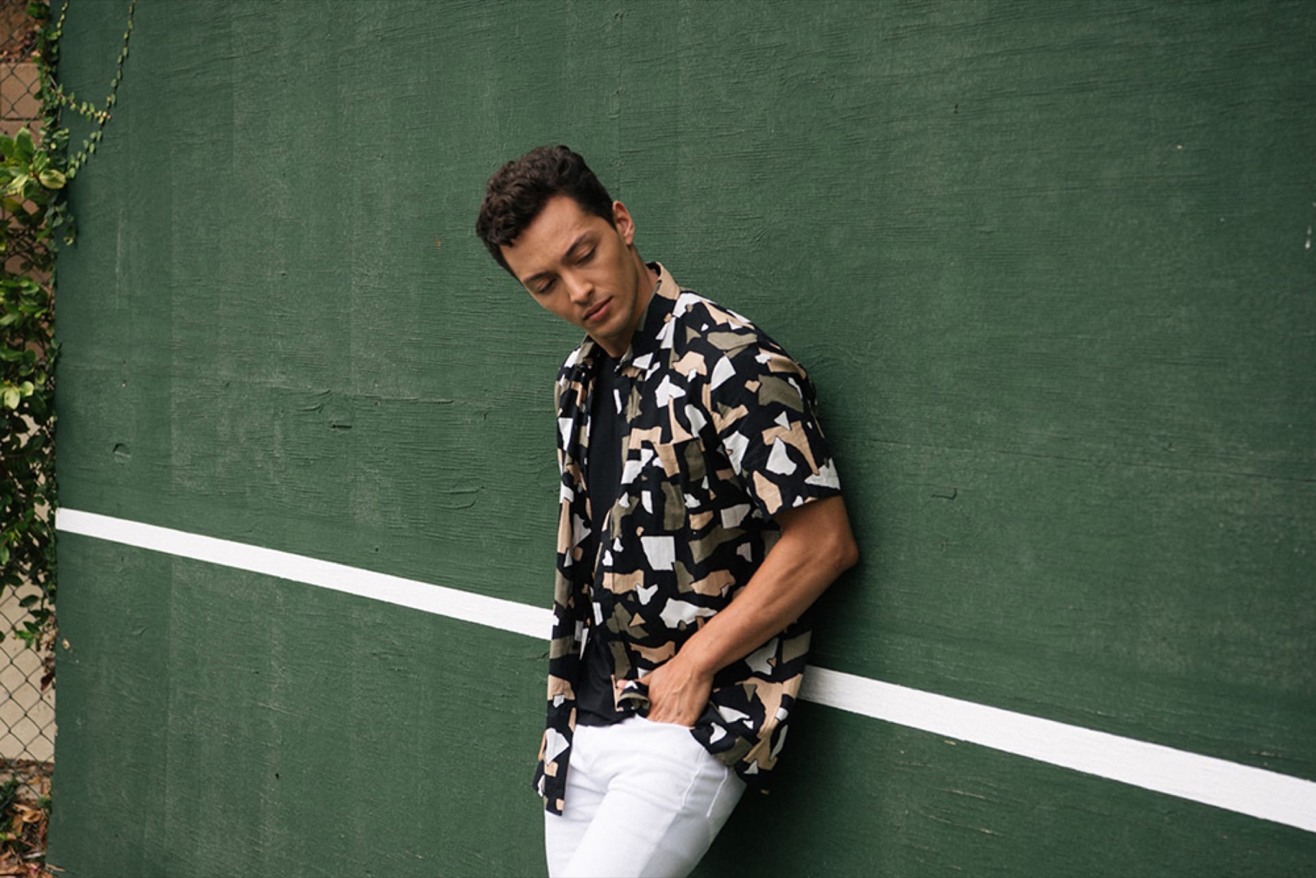 Man in a printed shirt leaning on a green court wall