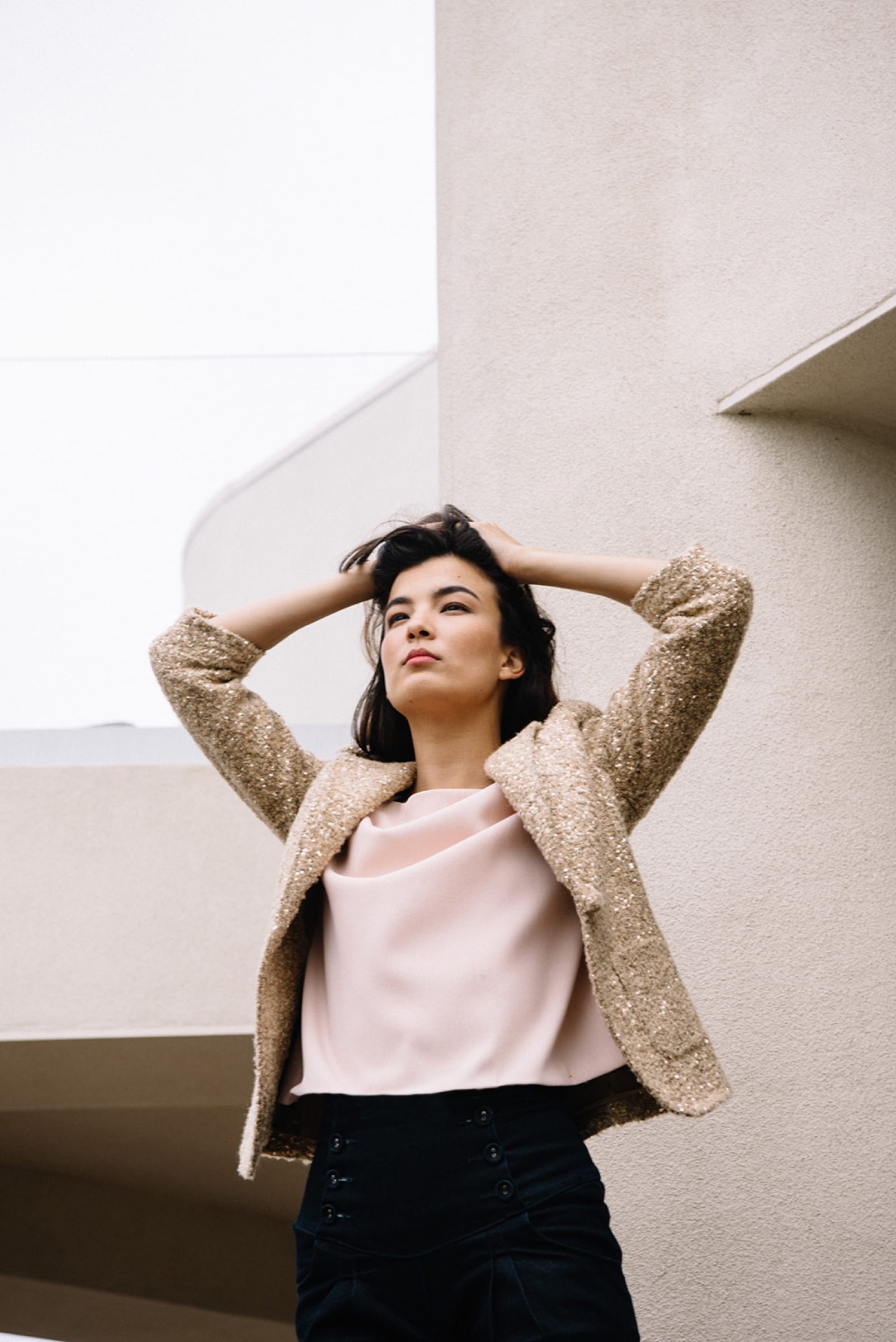 Woman in a beige jacket with hands in her hair by white architecture