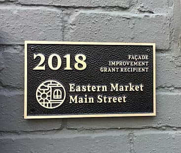 A bronze plaque on a brick wall: 2018 Façade Improvement Grant Recipient, with the Eastern Market Main Street medallion