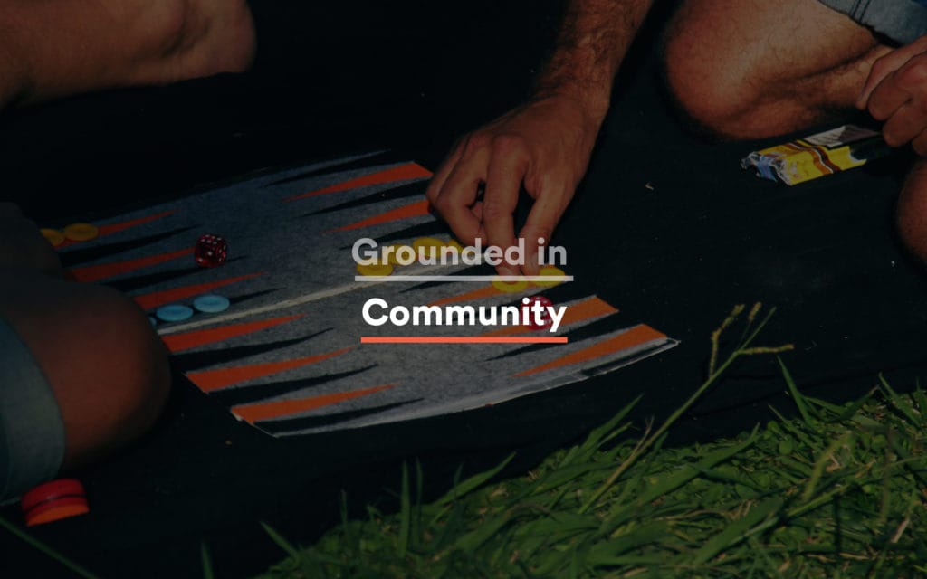Hands rolling dice in an outdoor backgammon game on a felt board laid on the grass, with "Grounded in Community" set in compressed sans white over the photograph