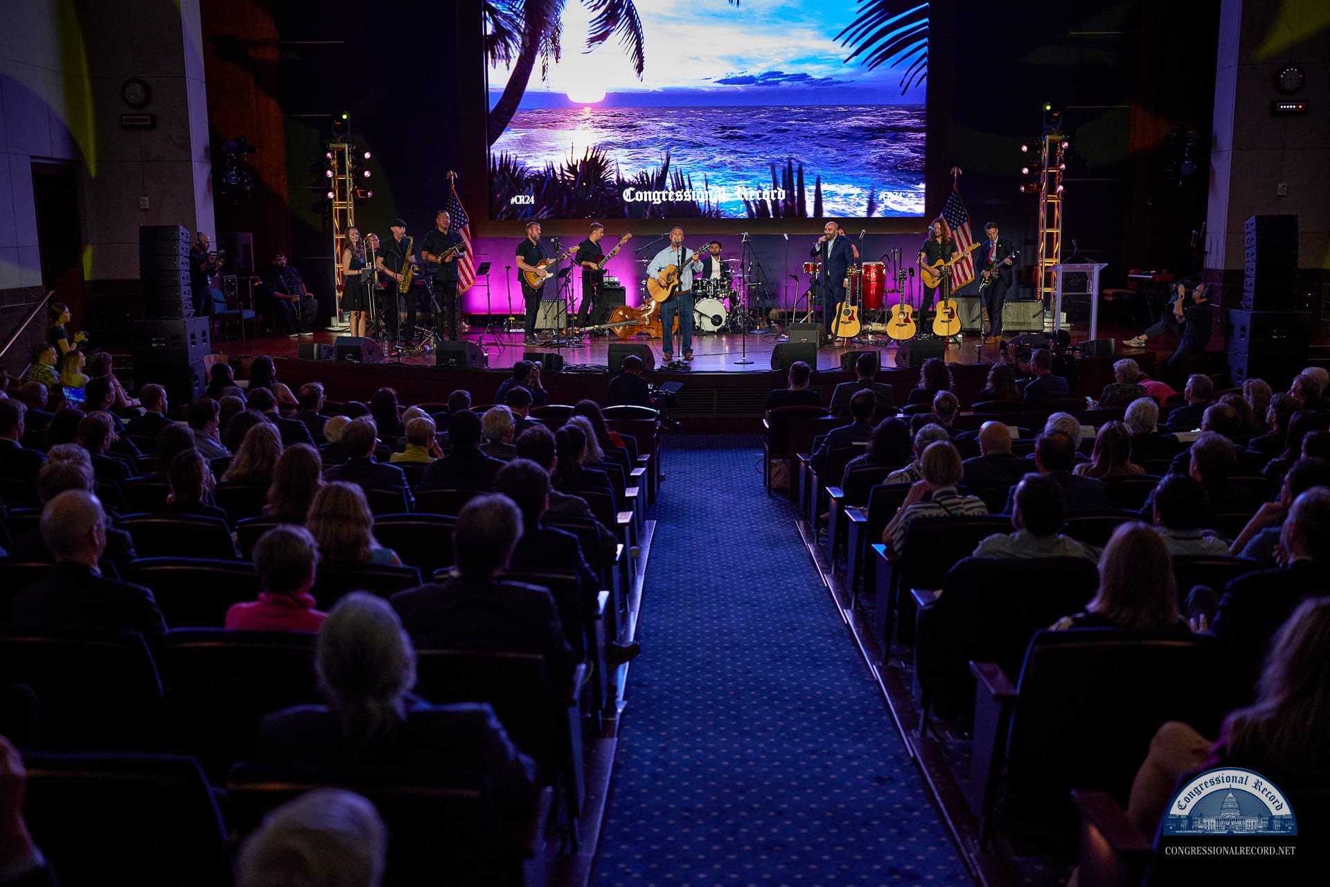 The room from the back: the full house and the band on stage under the palm-tree screen