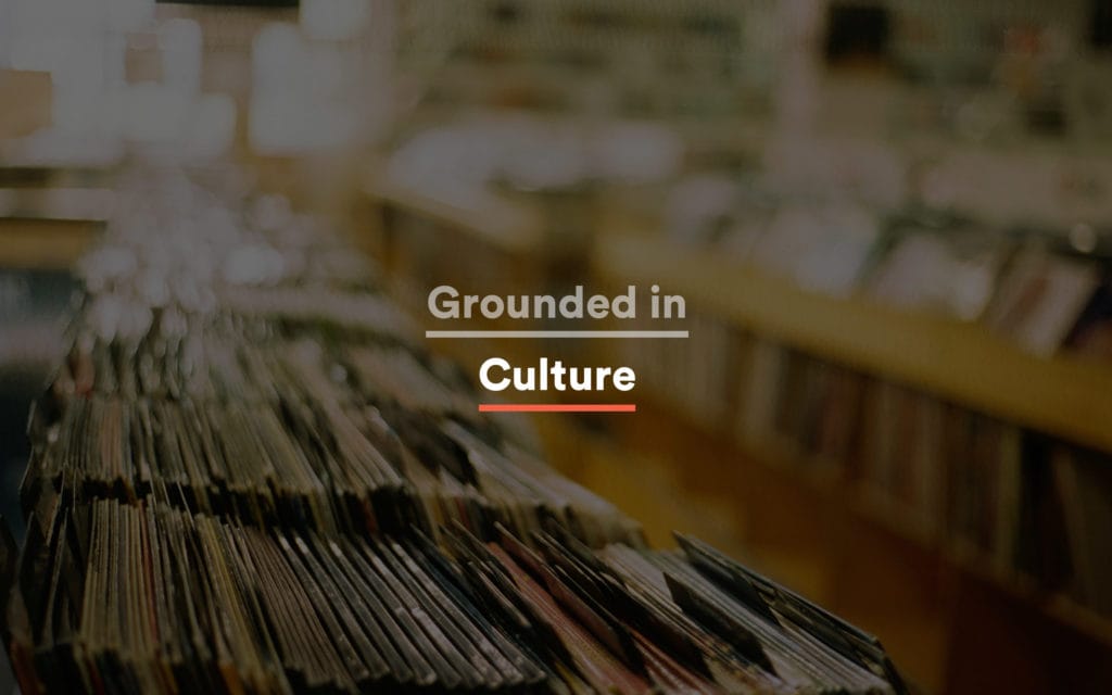 Brookland record store interior, vinyl shelves blurred at depth, with "Grounded in Culture" set in compressed sans white over the photograph