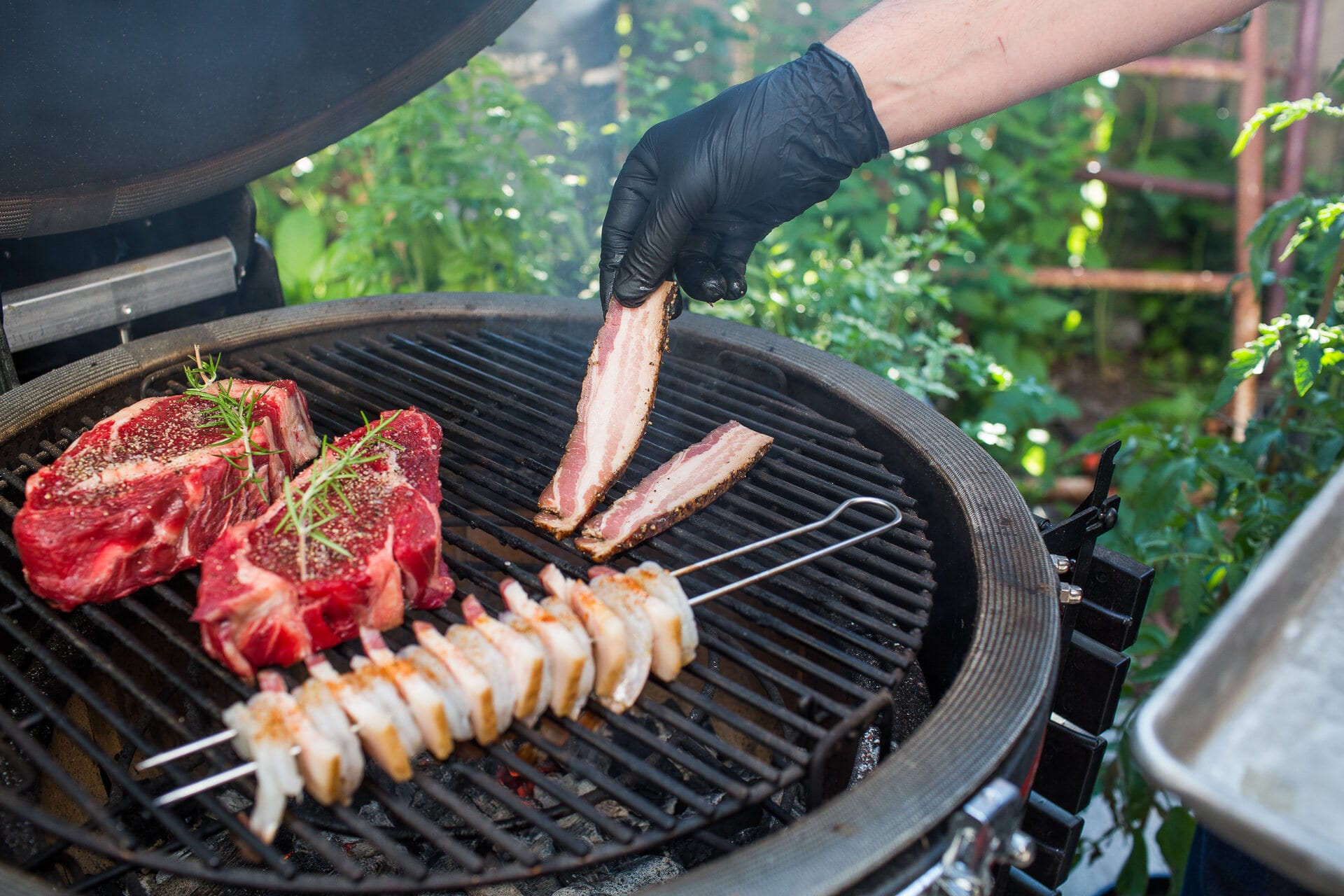 Gloved hand laying bacon on the grill over skewered pork