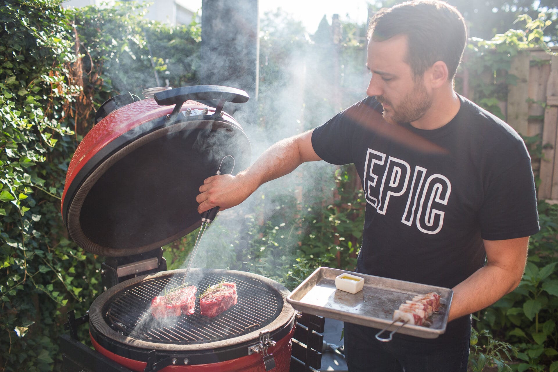 Cook in an EPIC tee lifting the lid of a red kamado grill, smoke rising