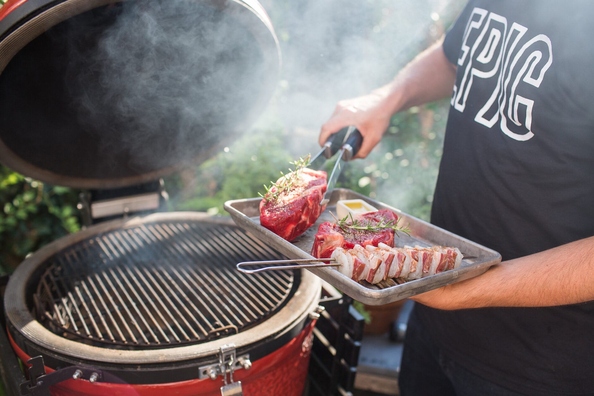 Cook in an EPIC tee carrying a tray of raw steaks past the kamado