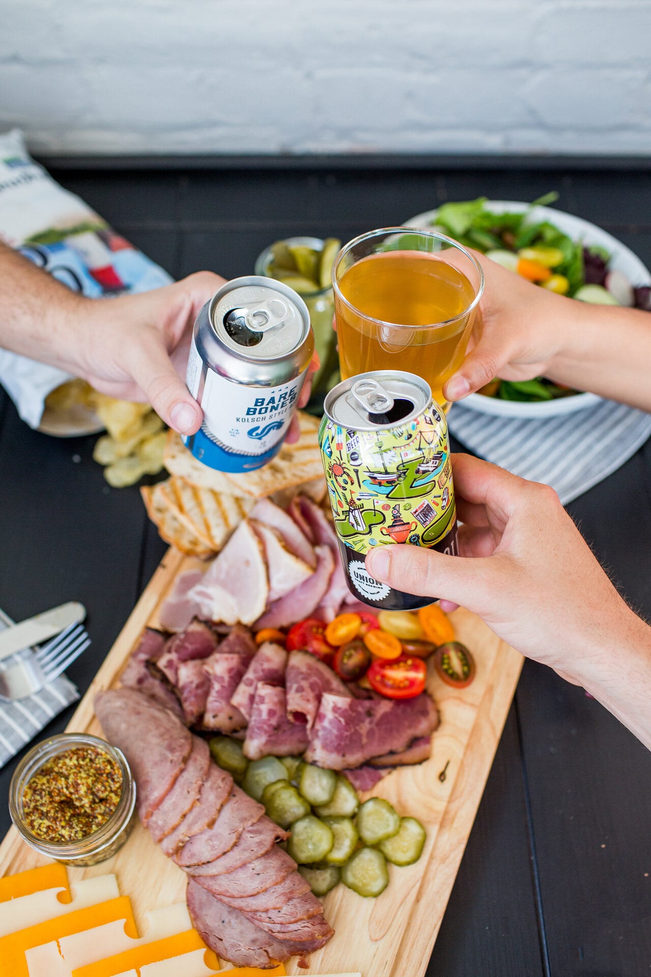 Hands toasting cans of beer over a charcuterie board