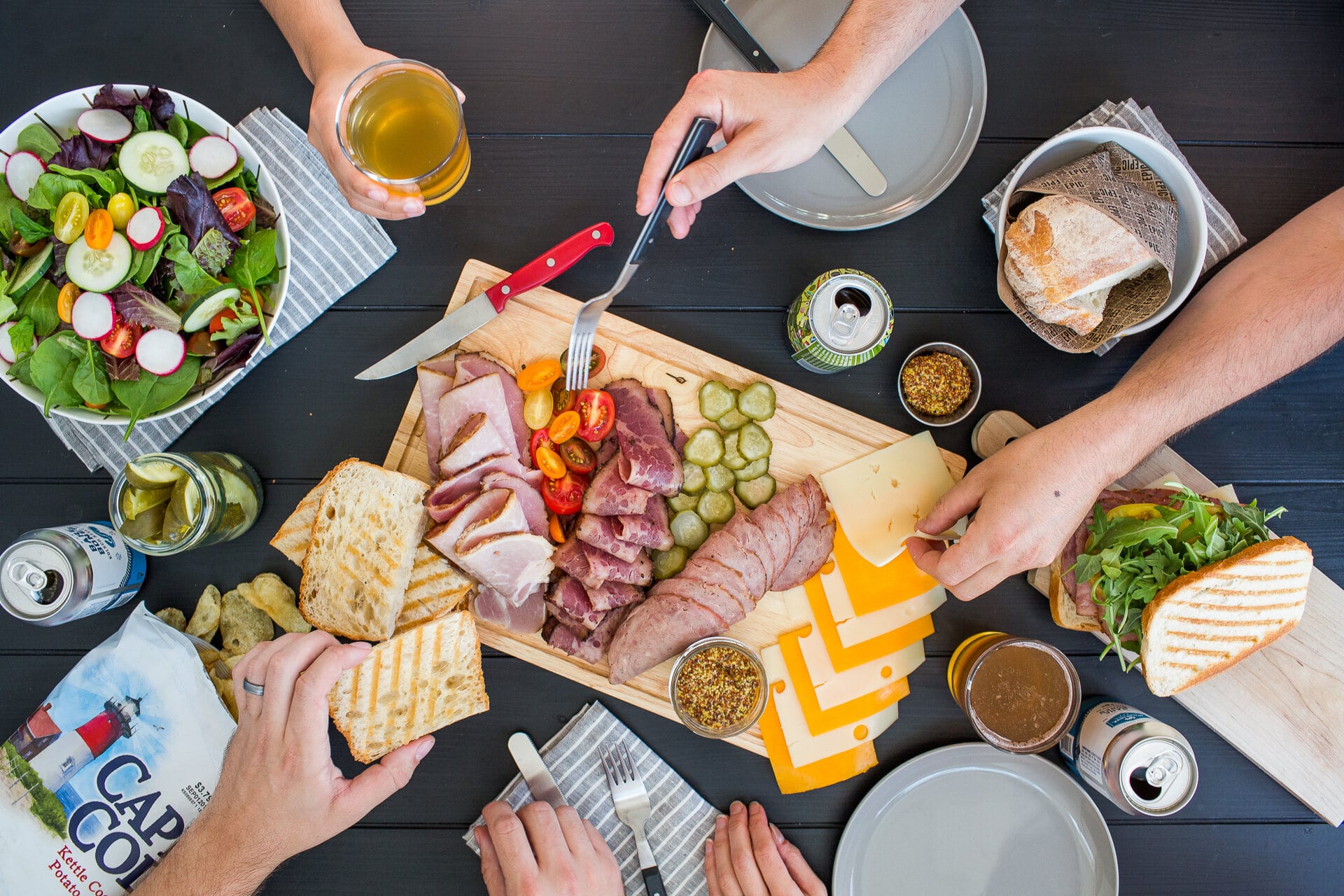 Overhead of hands cutting from a charcuterie board with a beer