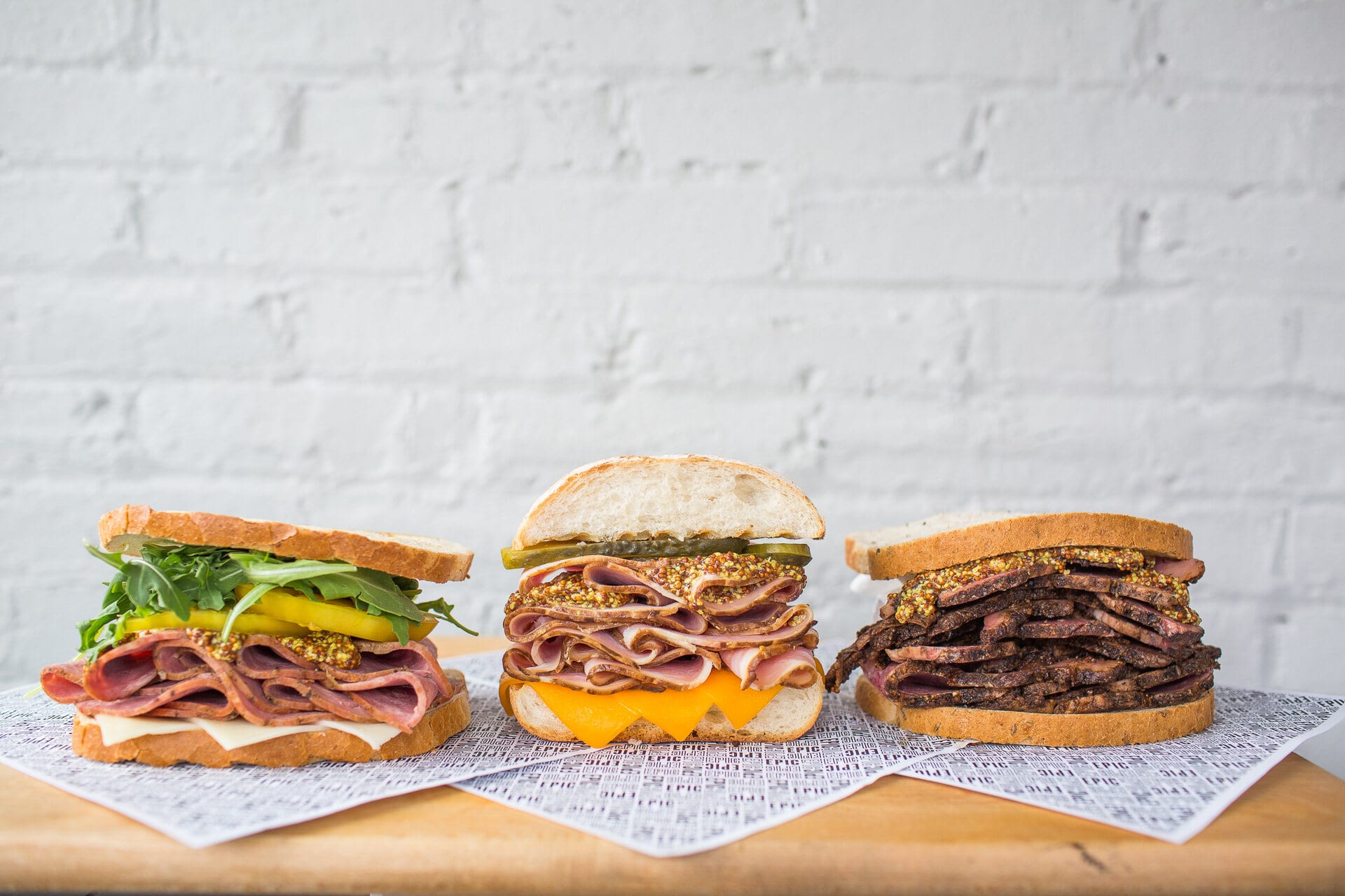 Three stacked deli sandwiches against a white brick wall